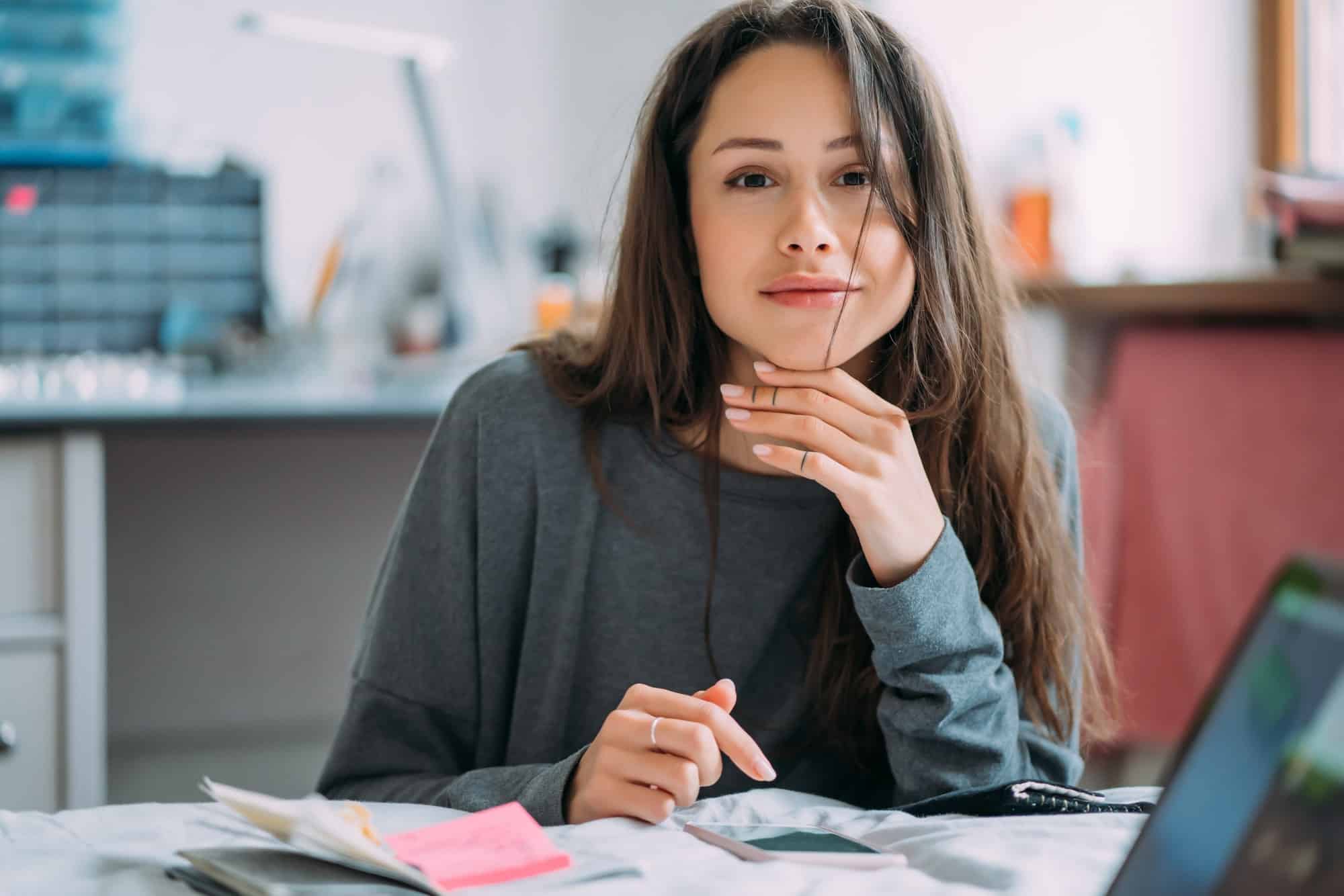 Portrait of smiling cute hipster girl staring on camera while writing college exercises in notebook